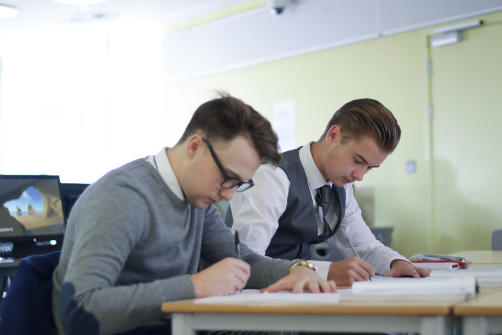 Two male Post 16 Longfield Academy students sit together whilst writing on paper with pens.