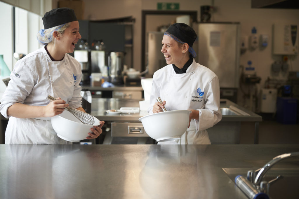 Two Longfield Academy students laughing together whilst whisking baking mixture in large plastic bowls.