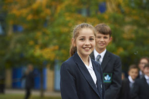 Two Longfield Academy students are pictured dressed in their academy uniform and smiling for the camera. A small group of students can be seen in the background behind them.