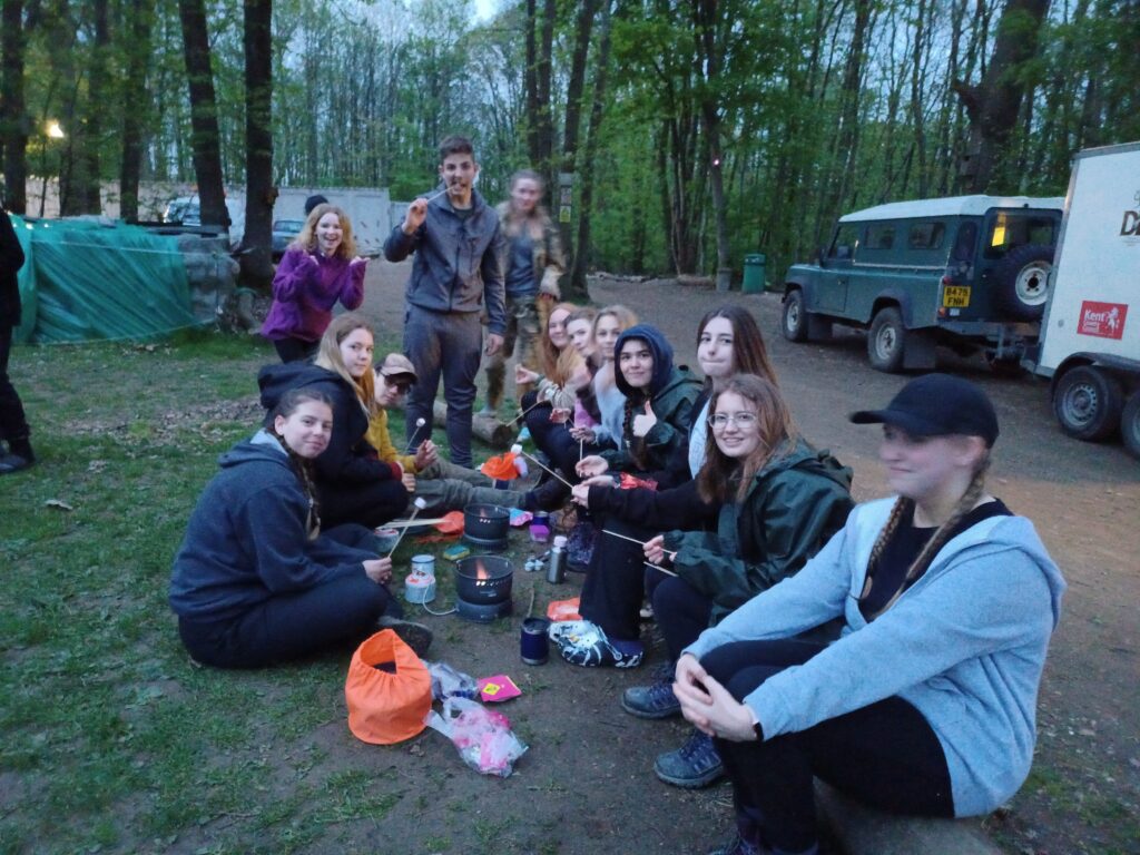 A large group of students are shown sitting together in a wooded area, toasting marshmallows and smiling for the camera.