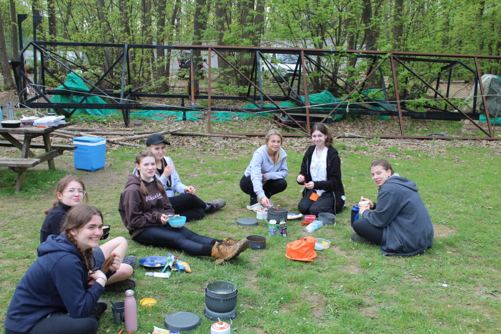 Seven students are seen sitting together in an outdoor wooded area, smiling for the camera whilst they eat.