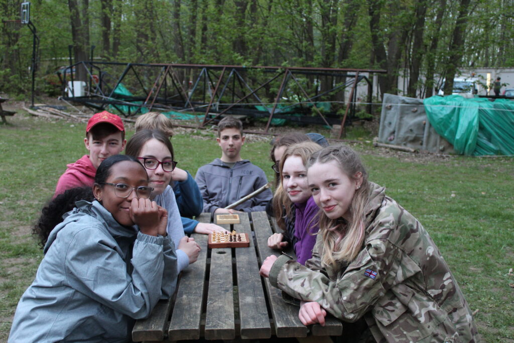 A group of students are seen sitting at a wooden table outdoors in a forest area, playing chess.