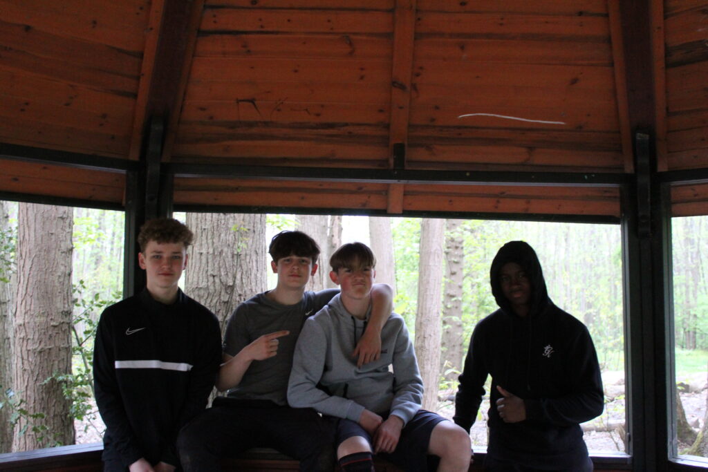 Four male students are seen posing for the camera whilst standing inside a wooden pavilion.