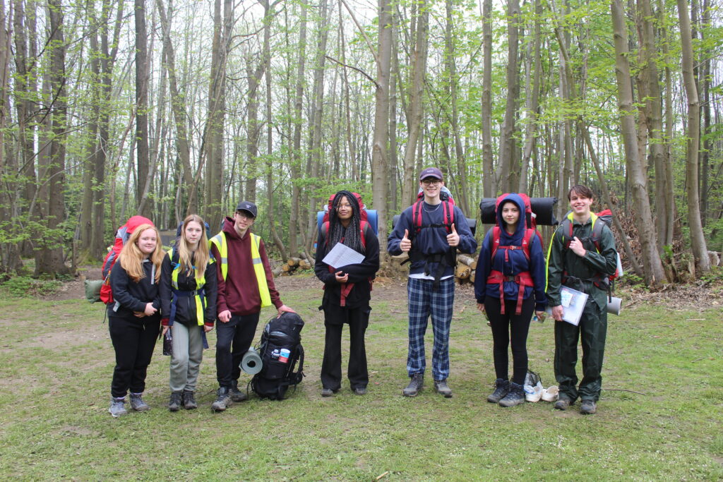 A group of seven students are pictured smiling for the camera, wearing their backpacks, whilst on a trek through the woods as part of their DofE practice event.