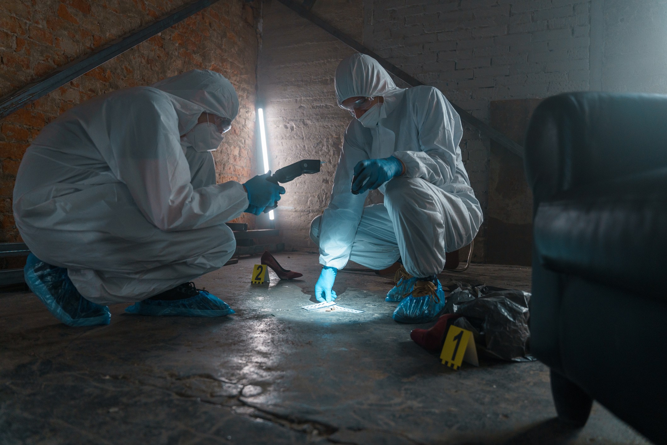 Criminologists in protective suits with camera taking photos of physical evidence in a flashlight light at the crime scene in a warehouse with brick walls