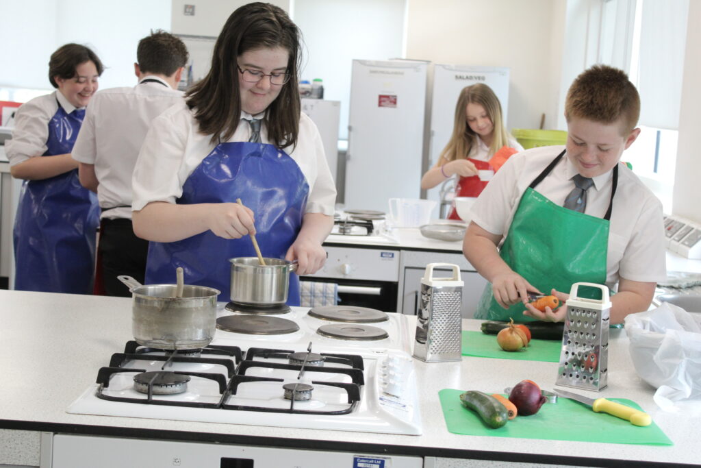 2 students cooking in a classroom, with one stirring in a pan, and another peeling a carrot