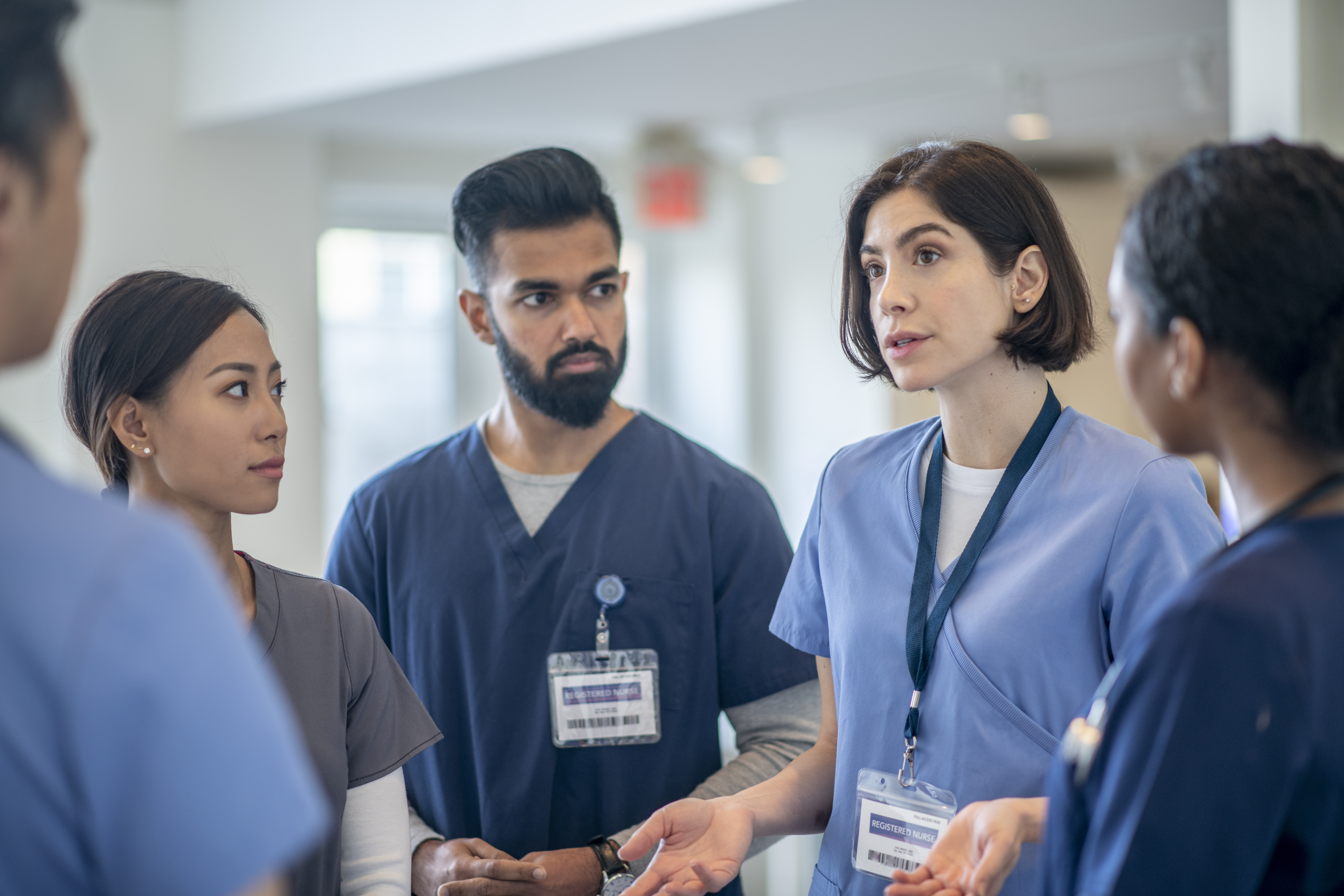 A small group of diverse medical professionals stand in the hallway for a brief meeting. They are each wearing scrubs and focused on the conversation.